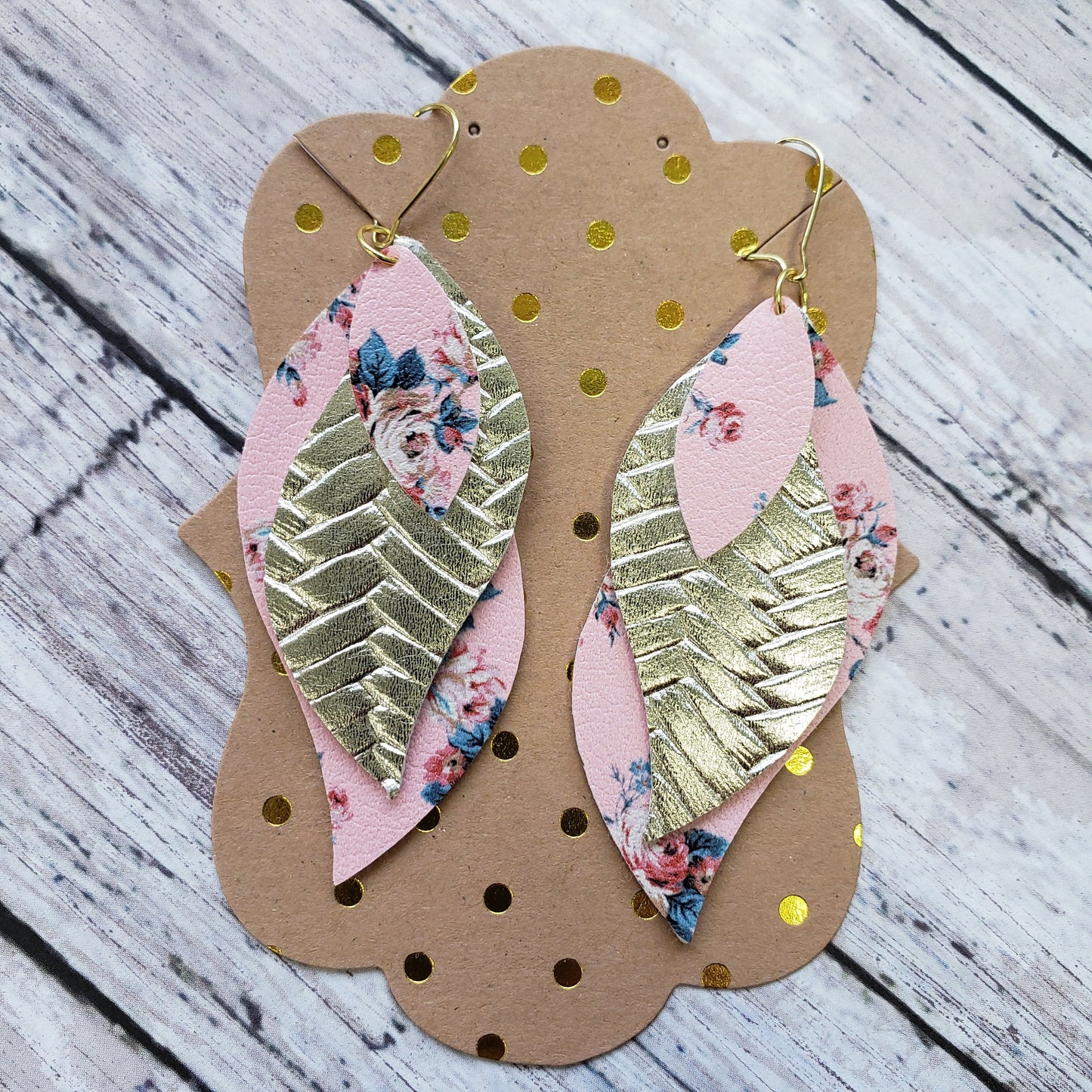 Triple layered faux leather earrings. Top layer pink with pink and blue floral print, middle layer pearlized metallic gold herring bone textures, bottom layer pink and blue floral print matching top layer on a gold french hook ear wire. Displayed on a kraft brown with metallic gold dot earring card.