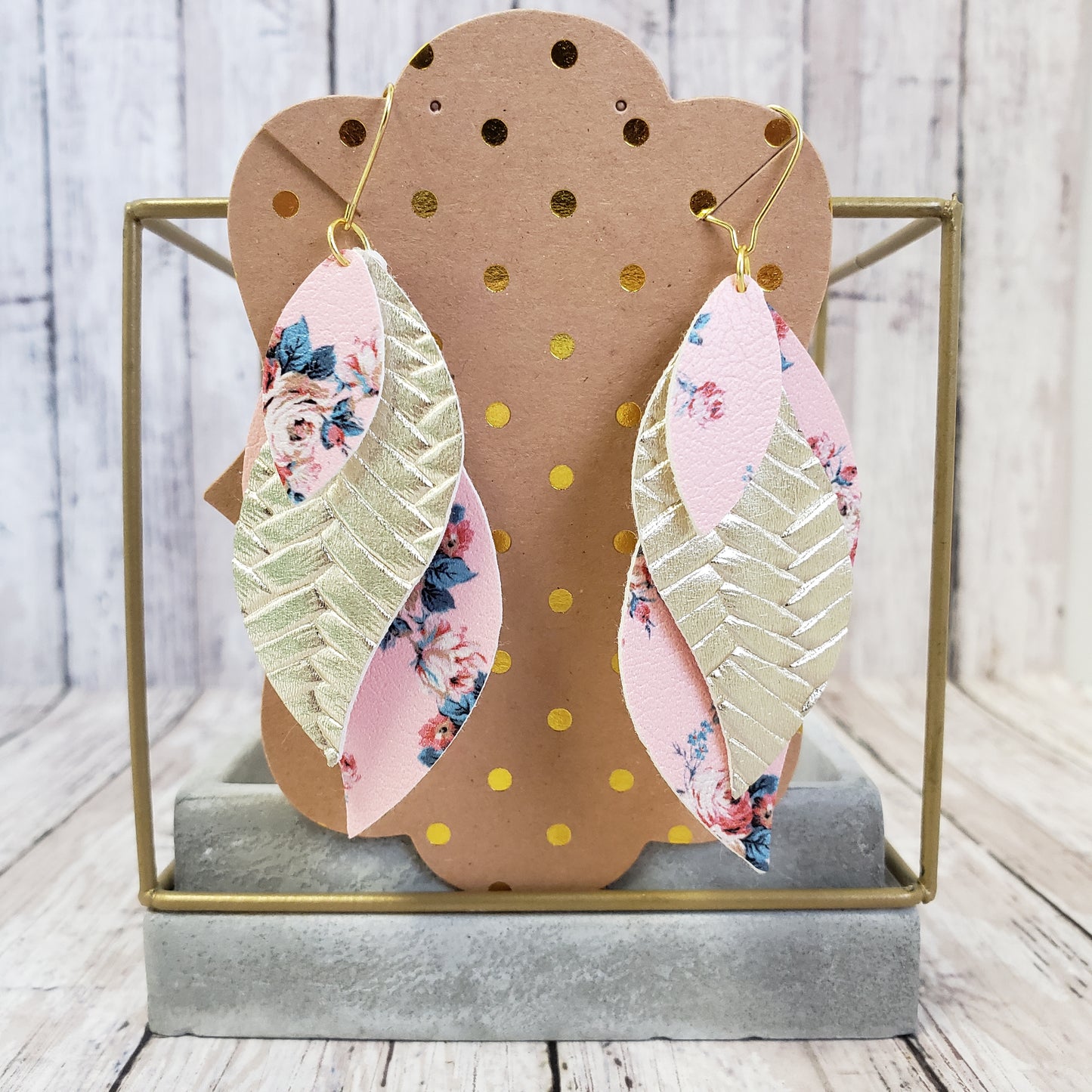 Triple layered faux leather earrings. Top layer pink with pink and blue floral print, middle layer pearlized metallic gold herring bone textures, bottom layer pink and blue floral print matching top layer on a gold french hook ear wire. Displayed on a kraft brown with metallic gold dot earring card.